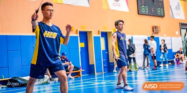 A male badminton player from Hangzhou International School standing on the sidelines of an indoor court, waiting to play.