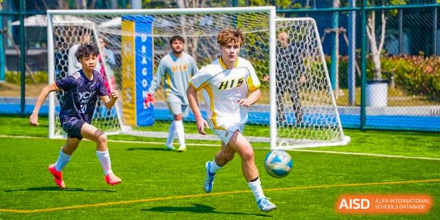 A male soccer player from Hangzhou International School in a white jersey dribbling the ball on an outdoor field during a game.