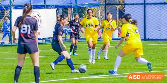 Female soccer players from Hangzhou International School in yellow and blue uniforms competing for the ball on a grass field.