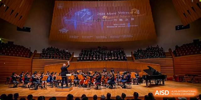 A large orchestra and choir performing on stage under a screen displaying the "Harrow Music Festival" banner, likely at Harrow LiDe School Chongqing (AISL Harrow Chongqing).