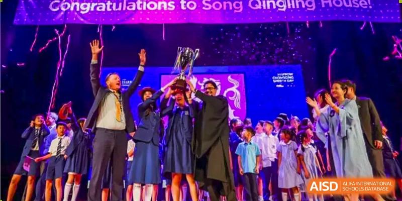 Students and staff from Harrow LiDe School Chongqing (AISL Harrow Chongqing) raising a large trophy on stage under bright lights, celebrating a victory.