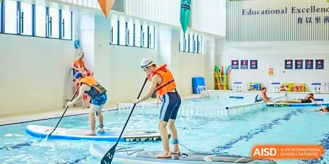 A student from Harrow LiDe School Chongqing (AISL Harrow Chongqing) wearing a life jacket and practicing stand-up paddleboarding in an indoor pool.