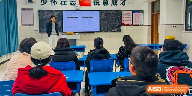 Students viewed from behind, sitting at desks and facing a teacher and a large screen displaying a presentation in a classroom Hefei No.6 High School International Department.