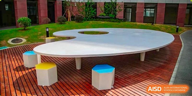 A modern outdoor seating area with a large round table and colored stools, showing the campus environment at the  Hefei No.6 High School International Department.