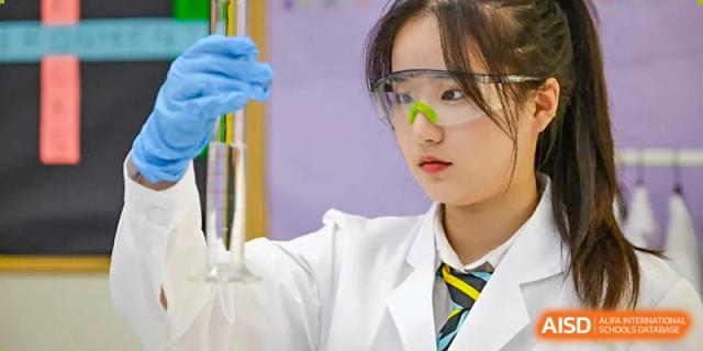 A female student from Hiba Academy Nantong in a lab coat and safety goggles, holding up a glass tube or graduated cylinder during a chemistry experiment.