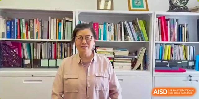 A female staff member or teacher from Hiba Academy Nantong, wearing glasses, smiling directly at the camera while standing in front of a large bookshelf filled with books.