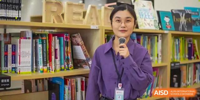 A female staff member or teacher from Hiba Academy Nantong speaking into a microphone while standing in front of a bookshelf, possibly during a school announcement or event.