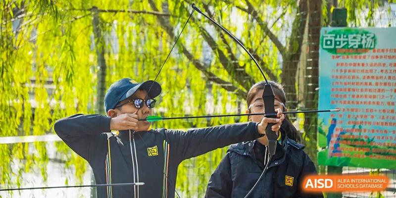 A Hiba Academy Nantong student wearing a hat and sunglasses aiming a bow and arrow during an archery lesson or activity.
