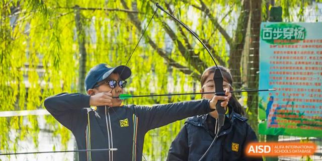 A Hiba Academy Nantong student wearing a hat and sunglasses aiming a bow and arrow during an archery lesson or activity.