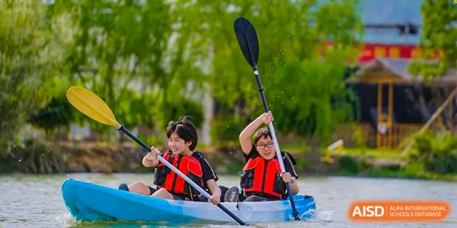 Two Hiba Academy Nantong students wearing life jackets, paddling a kayak or canoe on a green, tree-lined body of water.