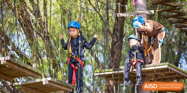 A student from Hiba Academy Nantong participating in a high-ropes or adventure course, wearing a helmet and harness while walking across a wooden bridge outdoors.