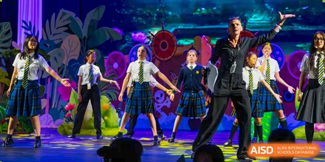 Students from Hiba Academy Nantong wearing school uniforms (plaid skirts and blazers) performing a choreographed dance on a brightly lit stage during a school production or event.