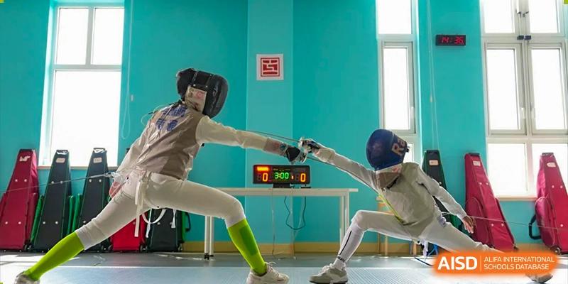 Two students from Hiba Academy Nantong engaging in a fencing match, both fully equipped in protective gear, with a digital scoreboard visible on a table between them.