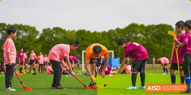 Four students from Hiba Academy Nantong playing a field sport like hockey or a similar stick-and-ball game on an outdoor grass field.