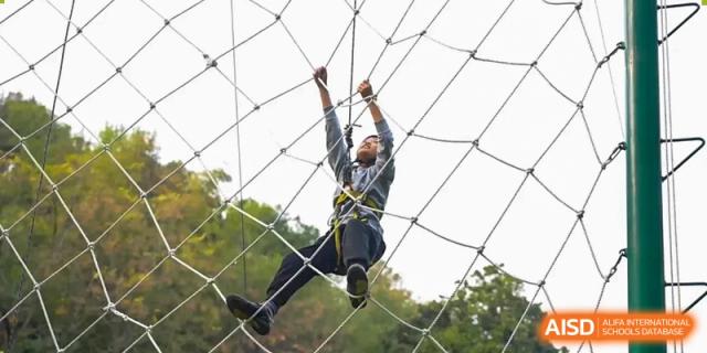 A student from Hiba Academy Nantong in a uniform climbing a large, knotted cargo net during an outdoor training or adventure activity.