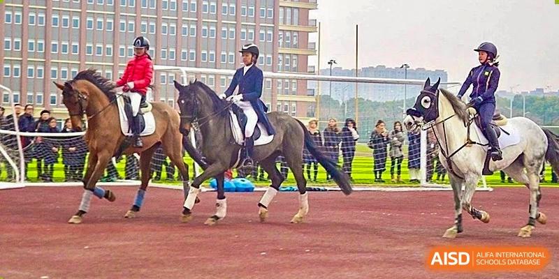 A student from Hiba Academy Nantong riding a dark horse in an equestrian competition or practice session on a red track, observed by others in the background.