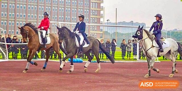 A student from Hiba Academy Nantong riding a dark horse in an equestrian competition or practice session on a red track, observed by others in the background.