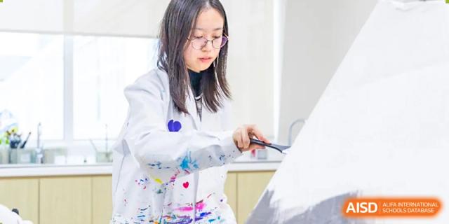 A female student from Hiba Academy Nantong, wearing a white lab coat, working with papers or a paintbrush in a brightly lit classroom or lab environment.