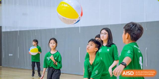 A group of students from Hiba Academy Nantong, dressed in green shirts, focused on a large, multi-colored beach ball during an indoor game or physical education class.