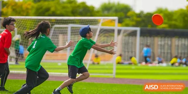 A young student from Hiba Academy Nantong, wearing a green shirt and a blue cap, running and reaching out to catch a ball on an outdoor sports field.