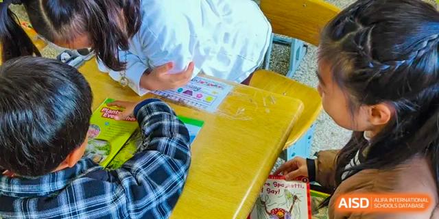 A close-up view of a teacher's or older student's hands pointing at a workbook or assignment on a student's desk at Kunming International Academy.