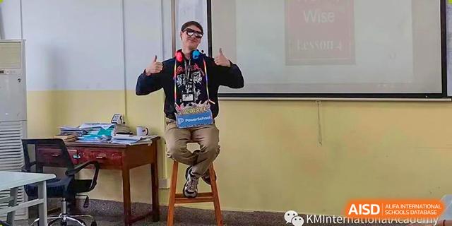A teacher wearing sunglasses and headphones, sitting on a stool and giving two thumbs-up in front of a projector screen during a presentation at Kunming International Academy.