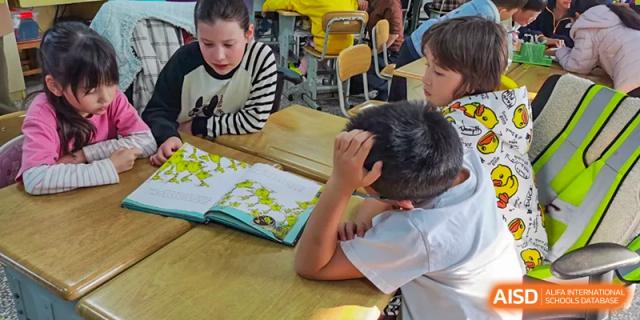 A young student resting his head on his hand while reading a textbook at a table with other students in a classroom at Kunming International Academy.