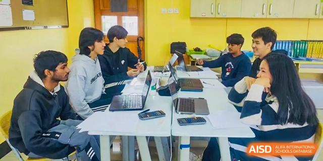 A group of older students collaborating on a project using laptops and tablets at a table in a brightly lit classroom at Kunming International Academy.