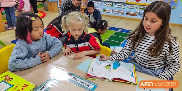 A young, blonde student sitting at a low table, reading a book with a teacher kneeling nearby in a primary classroom at Kunming International Academy.