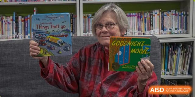 An older adult or teacher in a red plaid shirt holding up the children's book 'Goodnight Moon' in the school library at Kunming International Academy.