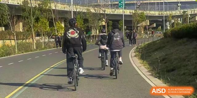 A group of students and faculty riding bicycles along a paved path next to a road, possibly for a field trip or outdoor activity sponsored by Kunming International Academy.