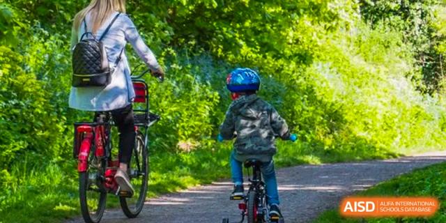 A young student wearing a helmet rides a bicycle down a shaded, tree-lined path, representing an outdoor activity sponsored by Kunming International Academy.