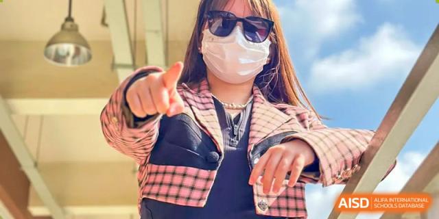 A student wearing sunglasses and a protective face mask, looking directly at the camera and pointing, possibly outside Kunming International Academy.