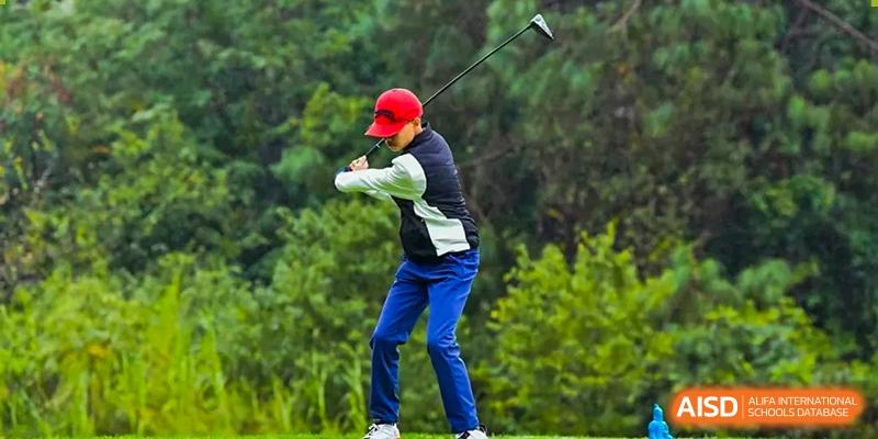 A student wearing a red cap and blue pants taking a swing during a golf lesson or practice session at Kunming International Academy.