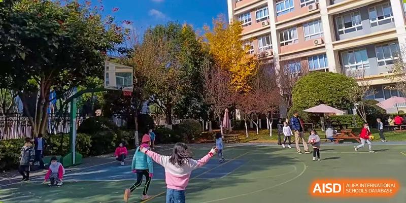 A female student spreading her arms wide and smiling while standing on an outdoor court with autumnal trees in the background at Kunming International Academy.