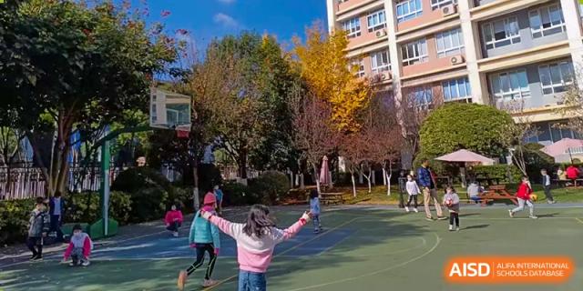 A female student spreading her arms wide and smiling while standing on an outdoor court with autumnal trees in the background at Kunming International Academy.