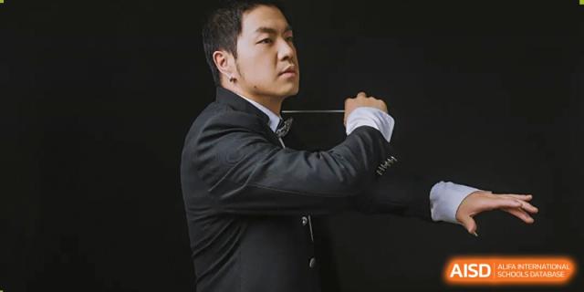 A conductor in a suit looking to the side while holding a baton, leading a musical performance at Kunming International Academy.