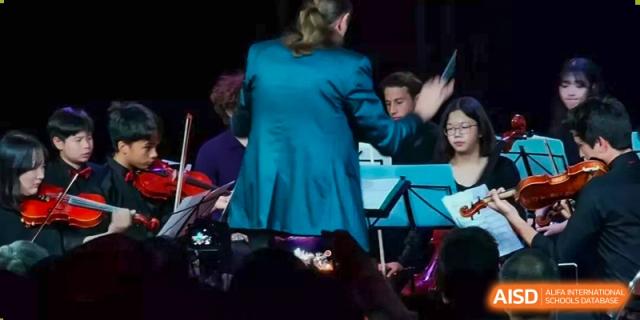A conductor in a teal jacket leading a group of musicians during an evening performance at Kunming International Academy.
