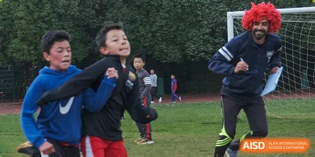 A young male student in a blue shirt running on a grassy field, possibly during a sports or recess activity at Kunming International Academy.