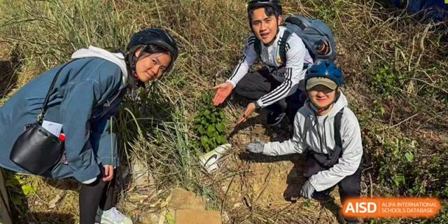 A small group of students and adults on an outdoor field trip or hiking excursion, pointing at plants or findings on the ground at Kunming International Academy.