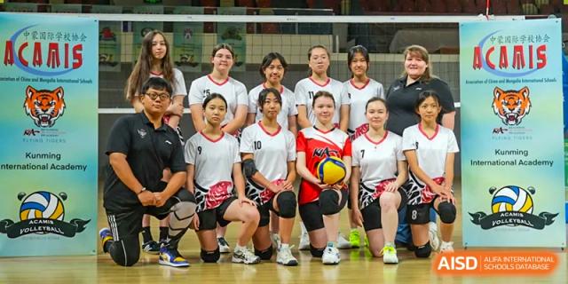 A women's volleyball team posing together for a group photo, some kneeling and one holding a ball, at Kunming International Academy.