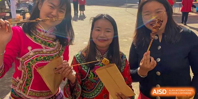 A female student in traditional clothing holding a craft or item with a smile, possibly at a cultural event at Kunming International Academy.