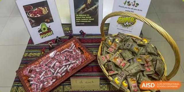 A basket filled with various packaged snack items and small figurines on a woven mat at a school fundraiser or market at Kunming International Academy.