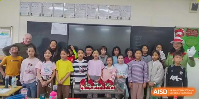 A group of female students posing with a cake decorated with red fruit in front of a whiteboard in a classroom at Kunming International Academy.