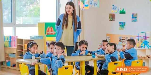 A female teacher interacting with a group of young primary students from Milton International School Qingdao who are seated at small desks in a classroom.