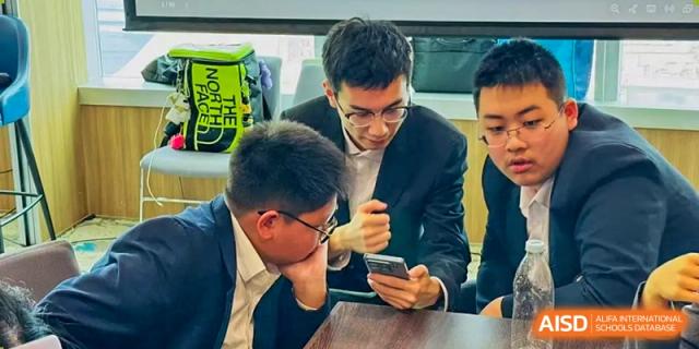 Students at Milton International School Qingdao collaborating on a school project, focused on a mobile device on a wooden desk.