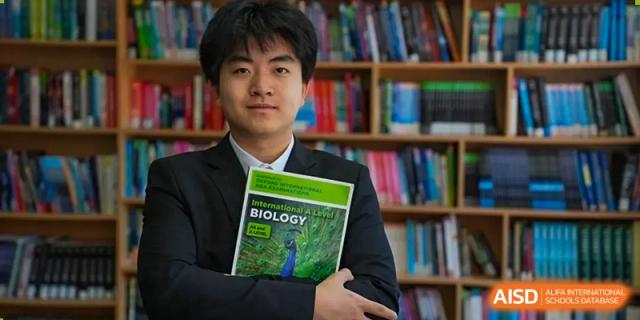 A male student from Milton International School Qingdao holding a 'Biology' textbook while standing in front of a well-stocked bookshelf in the library.