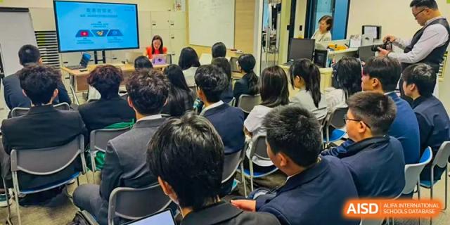 A wide shot of students from Milton International School Qingdao seated in an audience, attentively watching a presentation or lecture being given by a speaker at the front of the room.