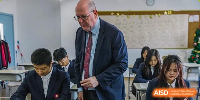 A male teacher or administrator from Milton International School Qingdao, wearing a suit and tie, interacting with students in a classroom.