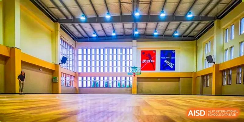 Interior view of an indoor gymnasium with wooden floors, bright overhead lights, and posters on the back wall at Milton International School Qingdao.